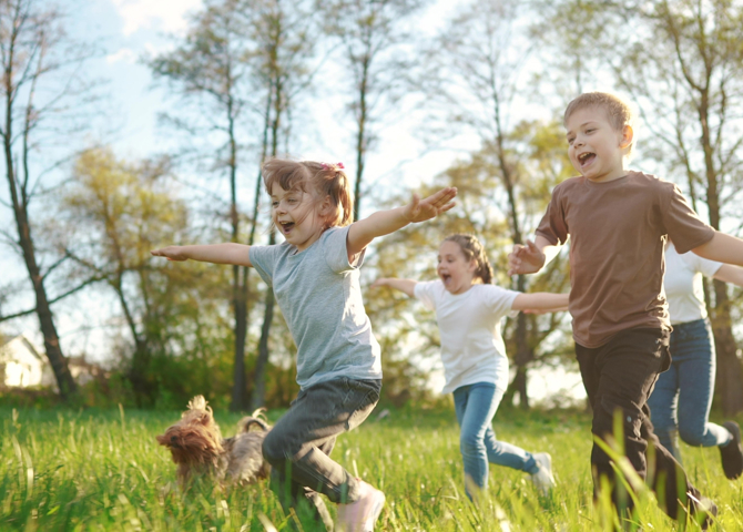 Güneşli günde çekilmiş fotoğrafta dört çocuk kollarını açmış, yanlarında kahverengi tüylü bir süs köpeğiyle yemyeşil çayırlarda mutlulukla koşuyorlar.