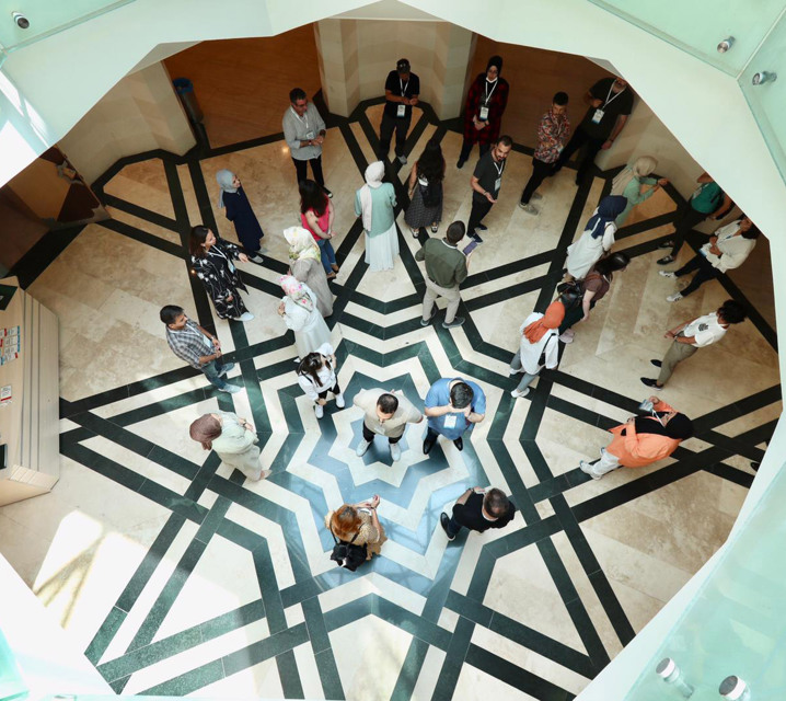 Yukarıdan çekilmiş bir fuaye alanının yer aldığı fotoğrafta; boyunlarına yaka kart takmış, bir grup genç kadın ve genç adam, iç içe geçmiş sekiz köşeli yıldız desenli zeminde dağınık bir halde ayakta duruyor.